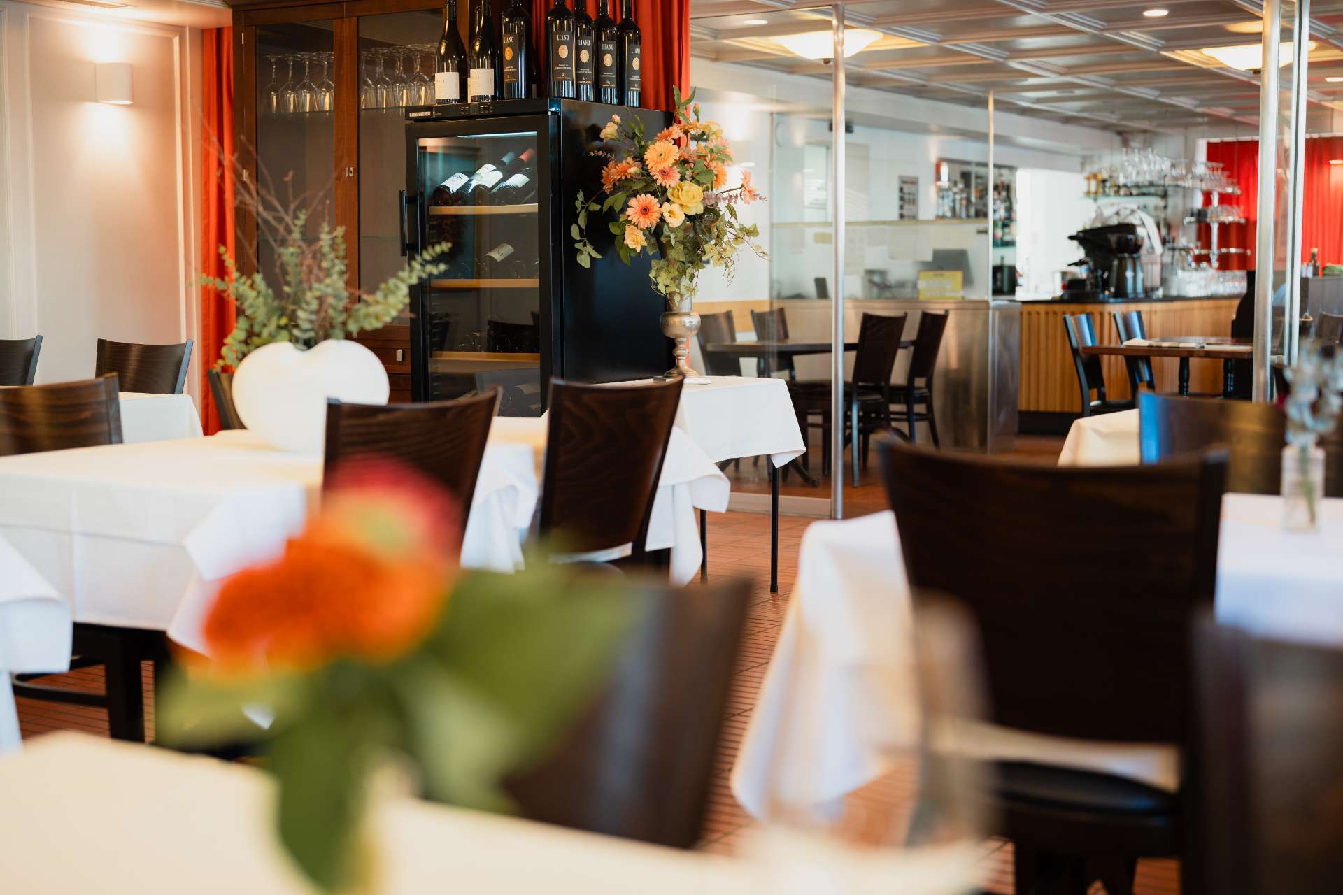 Restaurantinnenraum des Gasthof Hirschen mit weissen Tischdecken, dunklen Holzstühlen, Blumenschmuck, Weinschrank und Barbereich.